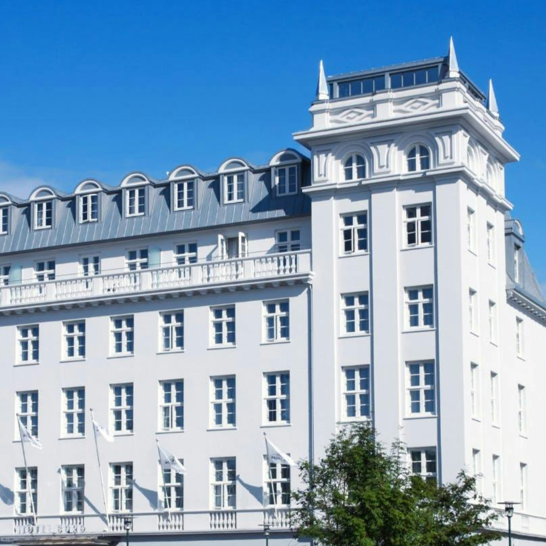 A captivating view of Hotel Borg in Reykjavik, Iceland, basking in the sunlight on a clear day. The elegant facade of the hotel stands against a vibrant blue sky, creating a striking contrast and highlighting the beauty of this iconic Icelandic landmark in the heart of the city