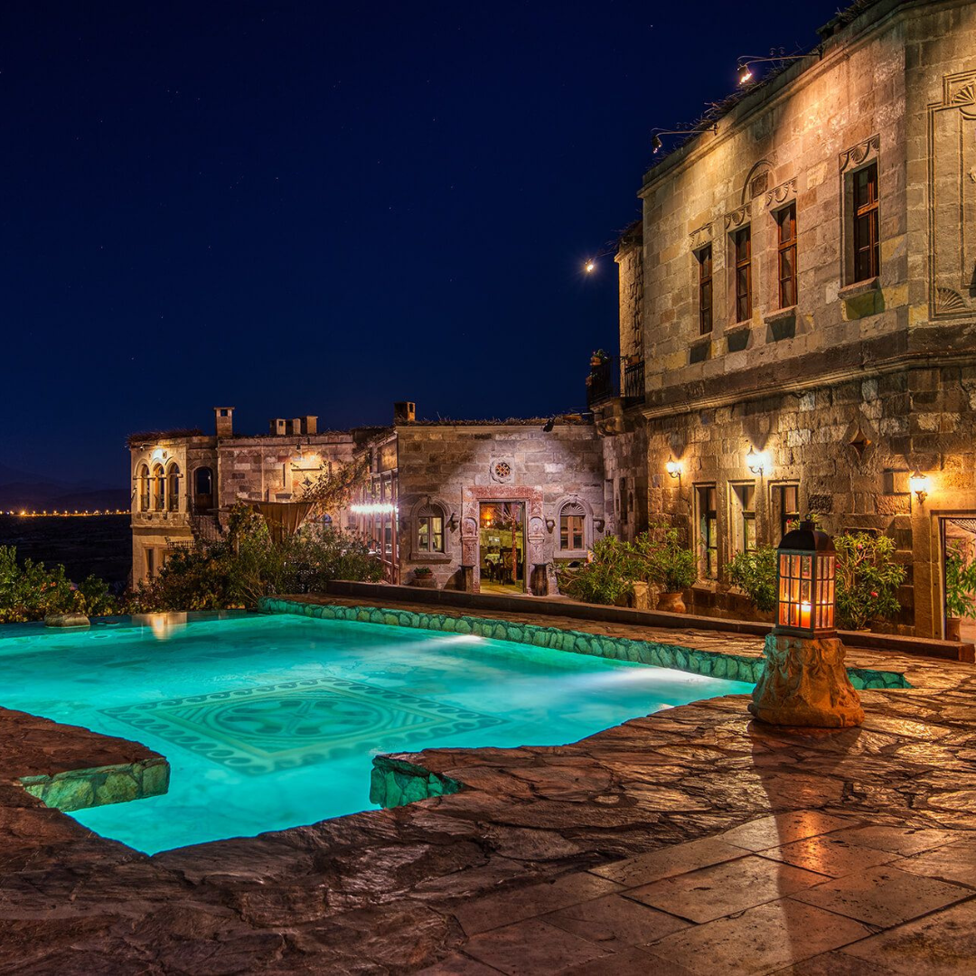 An enchanting evening view of the Museum Hotel in Cappadocia, Turkey. The pool takes center stage in the foreground, surrounded by the warm glow of twilight. In the background, the hotel's unique facade stands against the captivating Cappadocian landscape, creating a serene and luxurious atmosphere for guests.